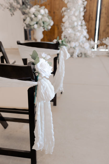 White rose and eucalyptus aisle chair flower rental with flowing white ribbon tie on dark wood folding chair at indoor barn wedding ceremony | Reno, Lake Tahoe & Sacramento