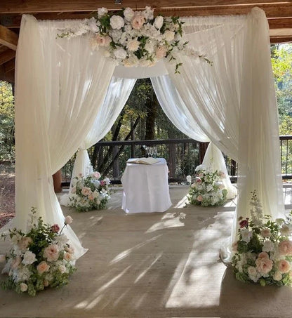 Full front view of chuppah rental with white chiffon drapery, Love Story arch adornment and aisle flowers in lush blush and white rose and peony floral, four Love Story aisle flowers at base corners, and white draped ceremony table at outdoor covered deck wedding venue with pine forest backdrop | Reno, Lake Tahoe & Sacramento