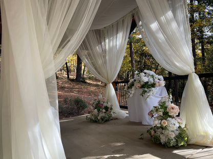 Interior view of chuppah rental with white chiffon drapery tied back, blush and white floral ground clusters at base corners, and white draped cocktail table with floral arrangement at outdoor pine forest wedding venue | Reno, Lake Tahoe & Sacramento