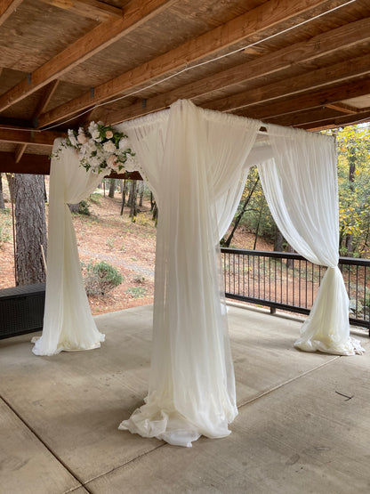 Full exterior view of chuppah rental with flowing white chiffon drapery pooling on ground and Love Story center arch adornment in white and blush floral at outdoor covered deck wedding venue with pine forest and fall foliage backdrop | Reno, Lake Tahoe & Sacramento
