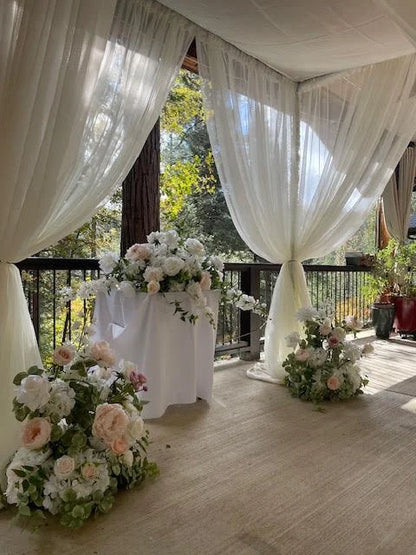 Full chuppah rental with white chiffon drapery, Love Story aisle flowers with lush blush and white rose and peony floral, floral ground clusters at base, and white draped cocktail table at outdoor covered deck wedding venue with pine forest backdrop | Reno, Lake Tahoe & Sacramento