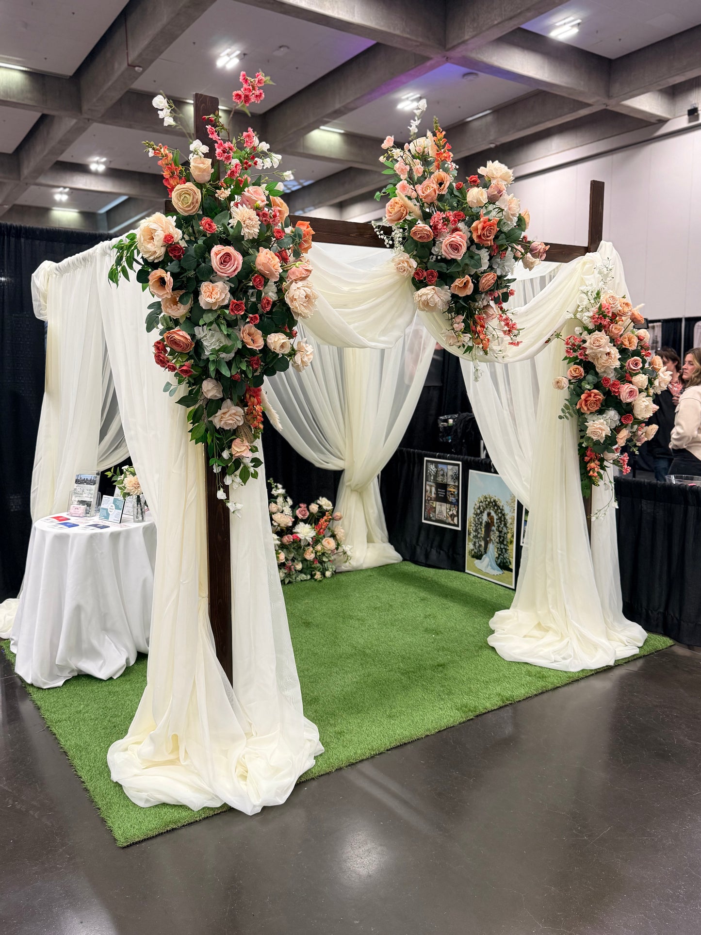 Timeless Arches bridal expo booth displaying chuppah rental with white chiffon drapery and Delicate arch adornments in coral, peach, and blush floral arrangements on top corners and base at indoor wedding bridal expo | Reno, Lake Tahoe & Sacramento