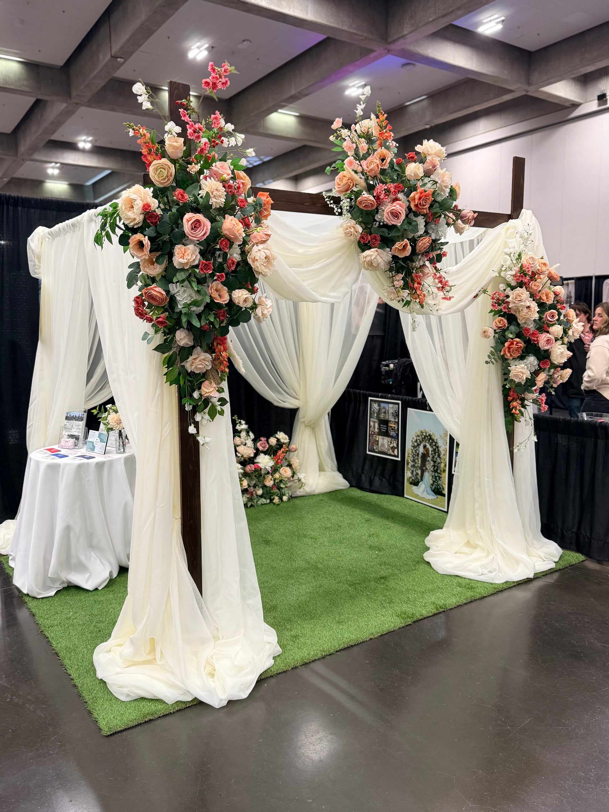 Timeless Arches bridal expo booth displaying chuppah rental with white chiffon drapery and Delicate arch adornments in coral, peach, and blush floral arrangements on top corners and base at indoor wedding bridal expo | Reno, Lake Tahoe & Sacramento