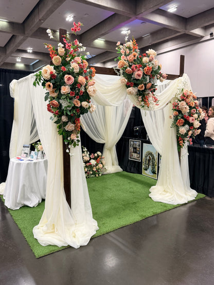Timeless Arches bridal expo booth displaying chuppah rental with white chiffon drapery and Delicate arch adornments in coral, peach, and blush floral arrangements on top corners and base at indoor wedding bridal expo | Reno, Lake Tahoe & Sacramento