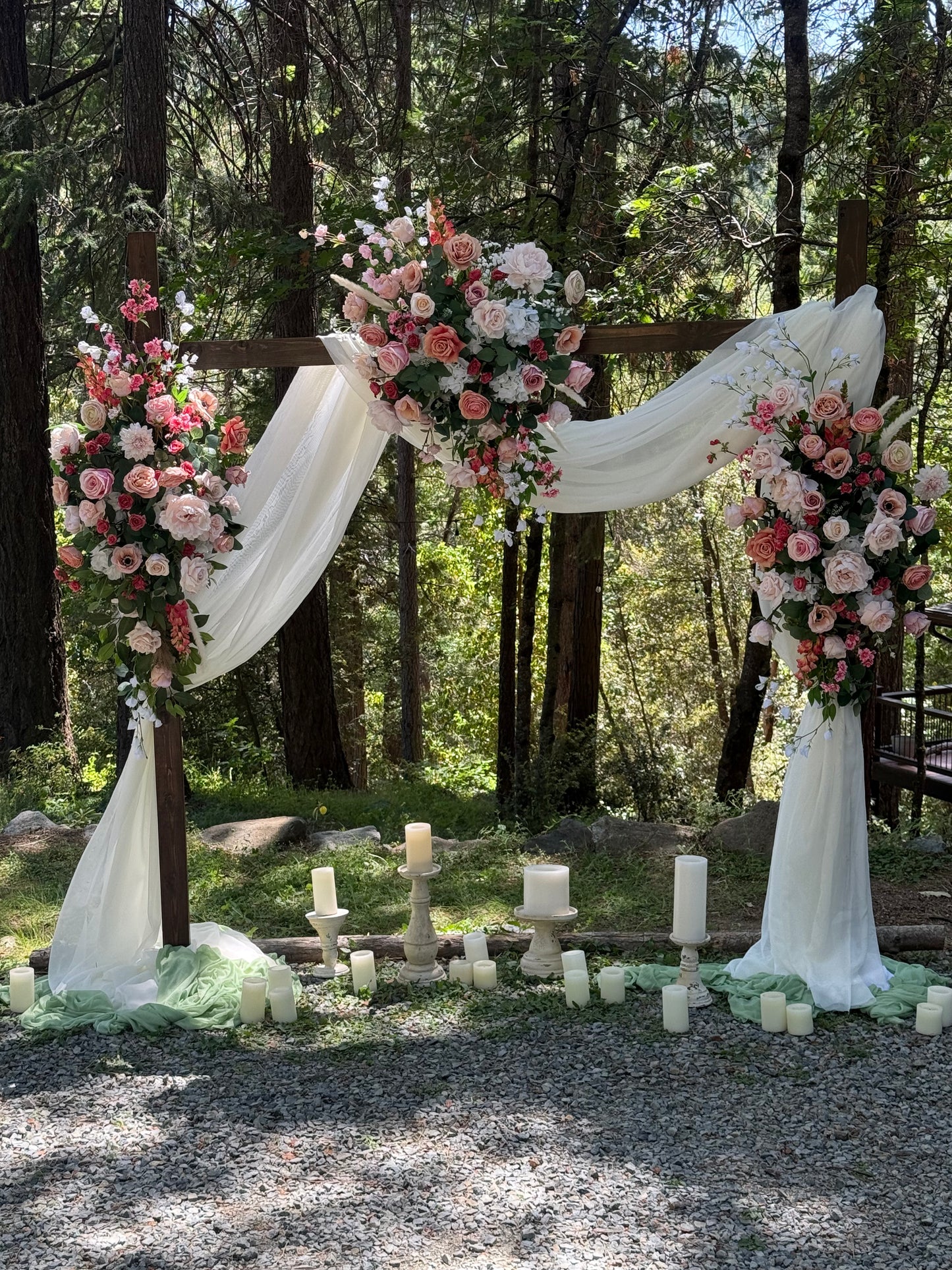 Full Delicate arch adornment rental with blush and coral roses, ranunculus, and white drapery on rustic wood arch at outdoor forest wedding with candles | Reno, Lake Tahoe & Sacramento