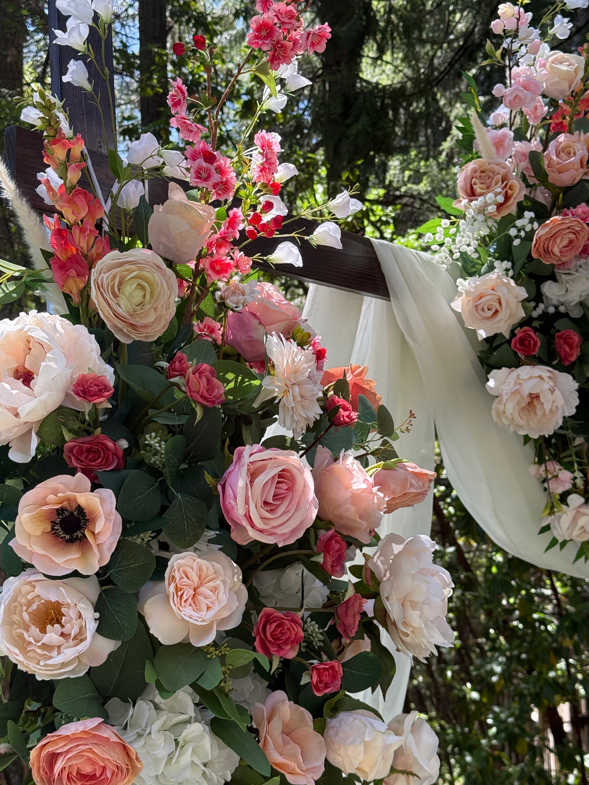 Close-up of Delicate arch adornment rental top corner with blush roses, snapdragons, ranunculus, and white drapery on rustic wood arch at outdoor forest wedding | Reno, Lake Tahoe & Sacramento
