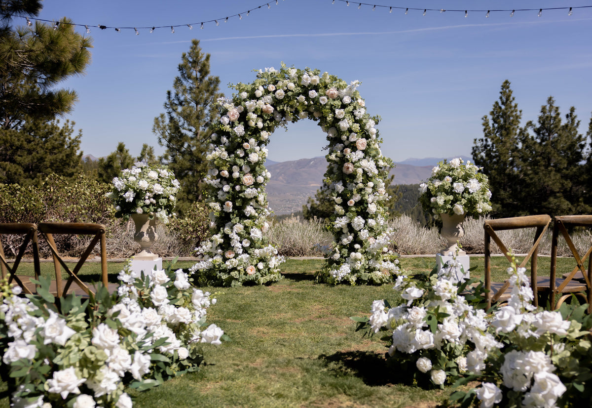 Enchanted floral arch rental with white roses, peonies, and greenery at outdoor Reno wedding ceremony with Enchanted aisle flowers and urns on white pedestals with mountain valley backdrop and white folding chairs | Reno, Lake Tahoe & Sacramento