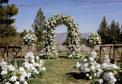 Enchanted floral arch rental with white roses, peonies, and greenery at outdoor Reno wedding ceremony with Enchanted aisle flowers and urns on white pedestals with mountain valley backdrop and white folding chairs | Reno, Lake Tahoe & Sacramento