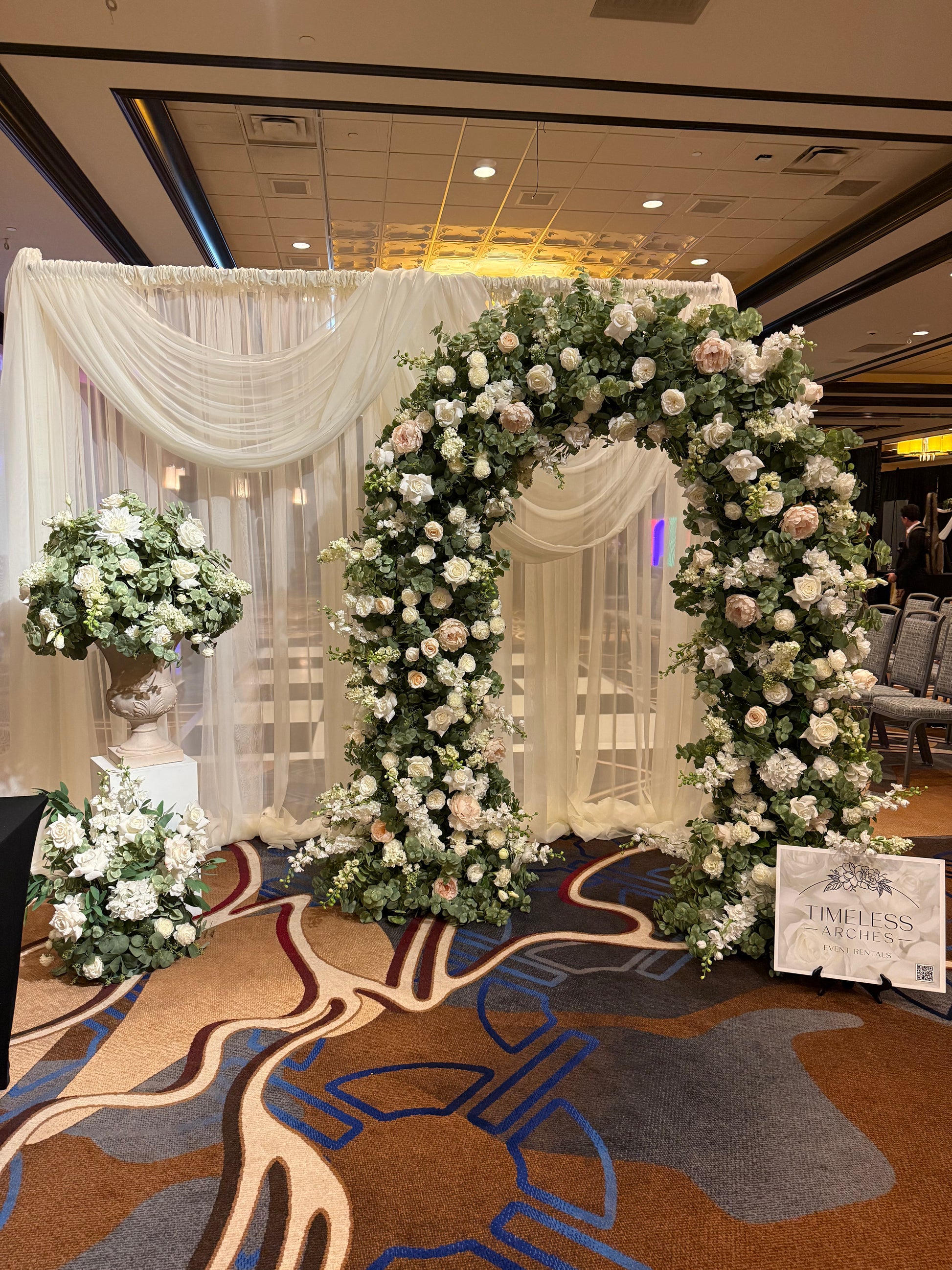 Timeless Arches bridal expo booth displaying Enchanted circular arch rental with blush and white roses and greenery, white draped backdrop, and Enchanted urn on white pedestal and single aisle flower| Reno, Lake Tahoe & Sacramento