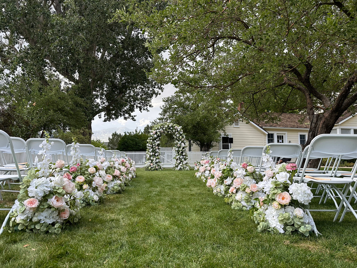 Wide aisle view of Enchanted floral arch rental at outdoor garden wedding ceremony with Love Story aisle flowers in blush and white, white folding chairs, and white picket fence backdrop | Reno, Lake Tahoe & Sacramento