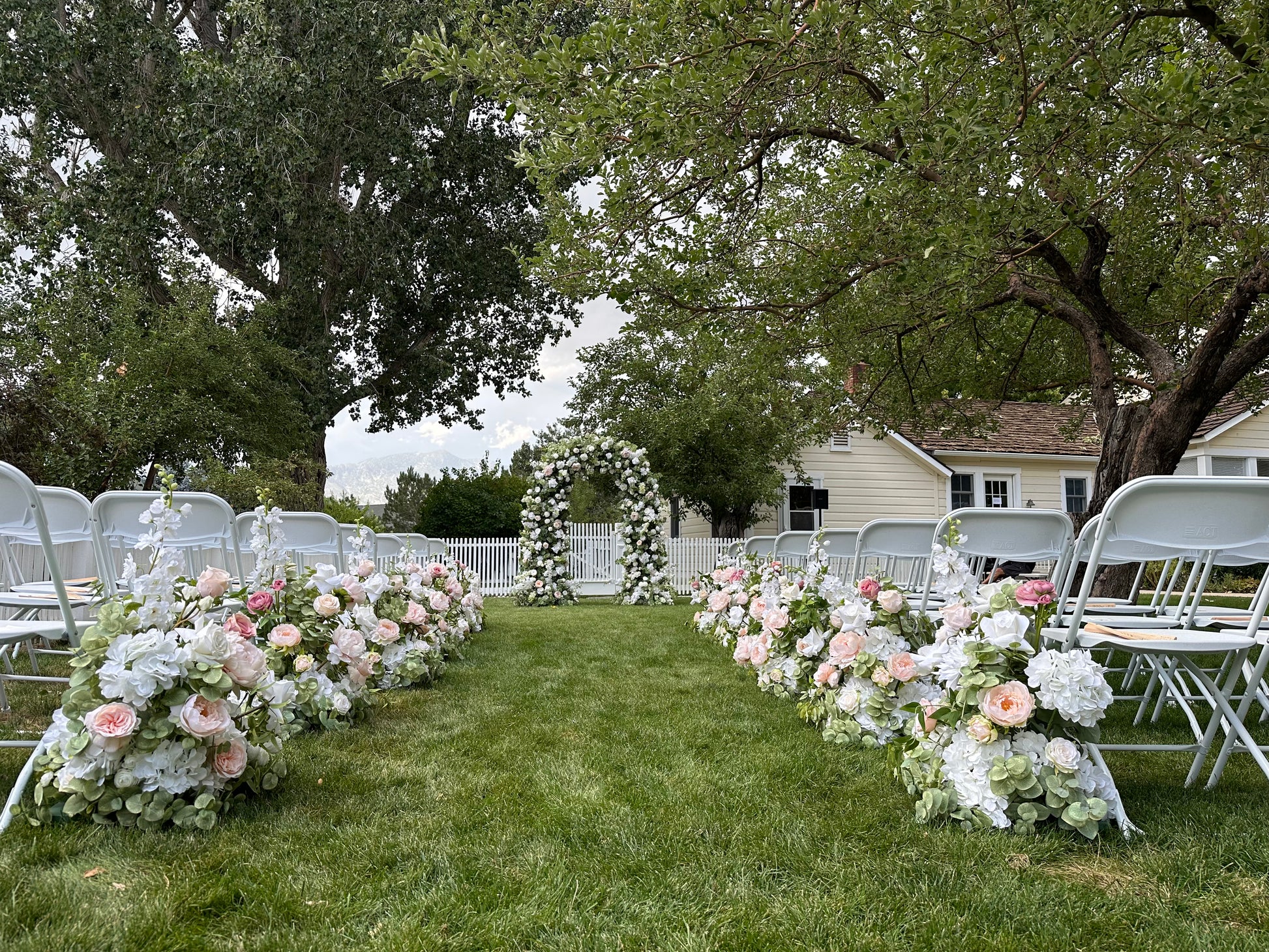 Wide aisle view of Enchanted floral arch rental at outdoor garden wedding ceremony with Love Story aisle flowers in blush and white, white folding chairs, and white picket fence backdrop | Reno, Lake Tahoe & Sacramento