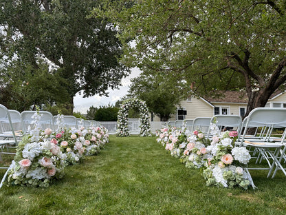 Wide aisle view of Enchanted floral arch rental at outdoor garden wedding ceremony with Love Story aisle flowers in blush and white, white folding chairs, and white picket fence backdrop | Reno, Lake Tahoe & Sacramento
