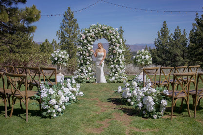 Bride standing under Enchanted circular arch rental with white roses, blush peonies, and greenery at outdoor Reno Nevada wedding venue with Enchanted aisle flowers and urns on 12" pedestals and mountain backdrop | Reno, Lake Tahoe & Sacramento