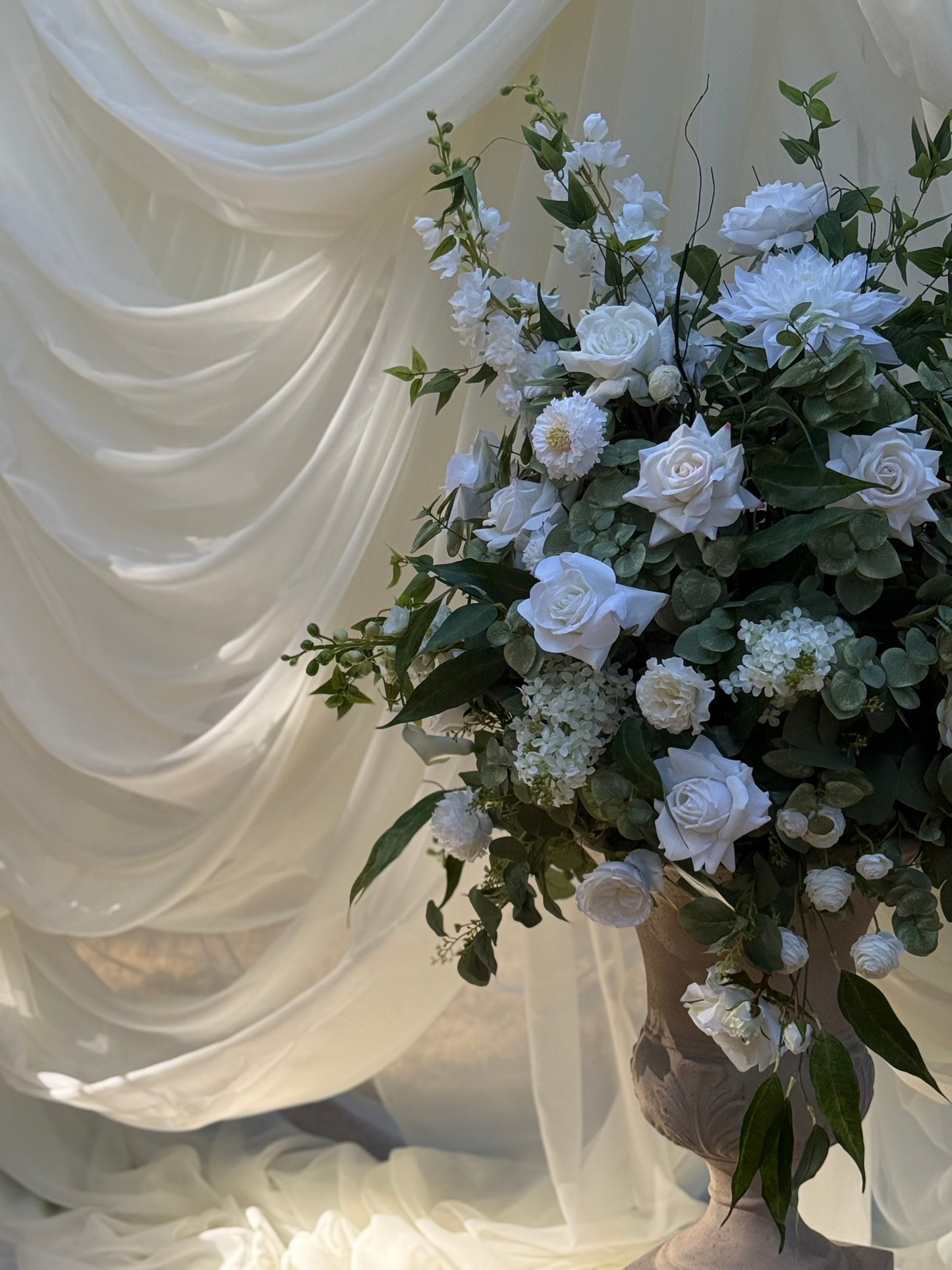 Close-up of Enchanted floral stone urn rental with white roses, white dahlias, white delphinium, white hydrangea, and lush eucalyptus against white draped backdrop at indoor wedding venue | Reno, Lake Tahoe & Sacramento