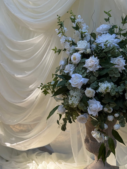 Close-up of Enchanted floral stone urn rental with white roses, white dahlias, white delphinium, white hydrangea, and lush eucalyptus against white draped backdrop at indoor wedding venue | Reno, Lake Tahoe & Sacramento