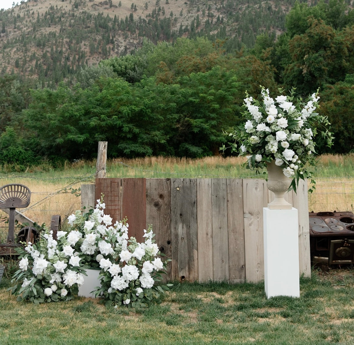 Enchanted aisle flower and urn rental on white square pedestal with white roses, white delphinium, and lush greenery at rustic outdoor wedding venue with reclaimed wood fence and mountain backdrop | Reno, Lake Tahoe & Sacramento