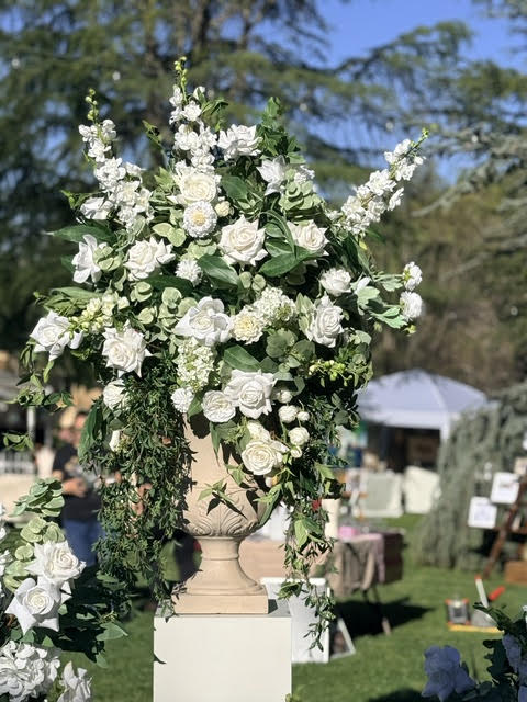 Enchanted floral stone urn rental on white pedestal overflowing with white roses, white delphinium, white hydrangea, and cascading greenery at outdoor wedding bridal market | Reno, Lake Tahoe & Sacramento