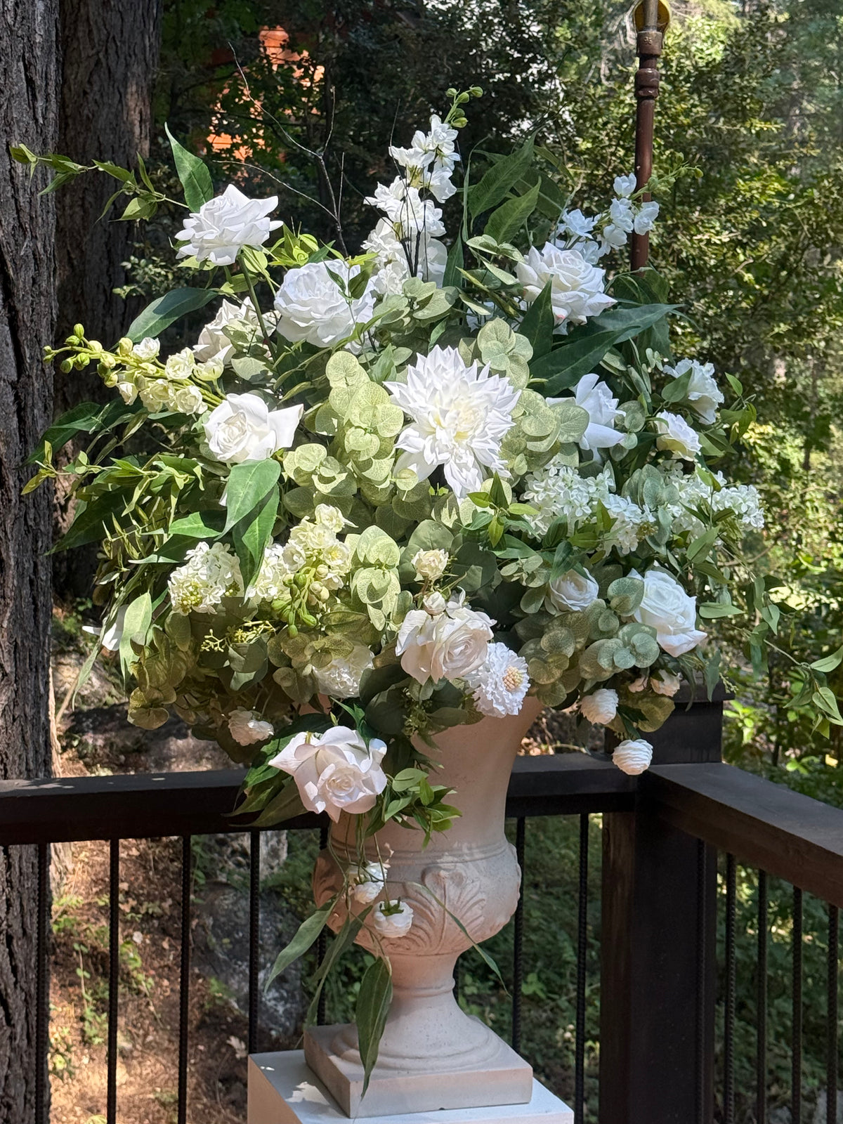 Enchanted floral stone urn rental on wood deck railing overflowing with white roses, white dahlias, white hydrangea, white delphinium, and lush greenery at outdoor pine forest wedding venue | Reno, Lake Tahoe & Sacramento