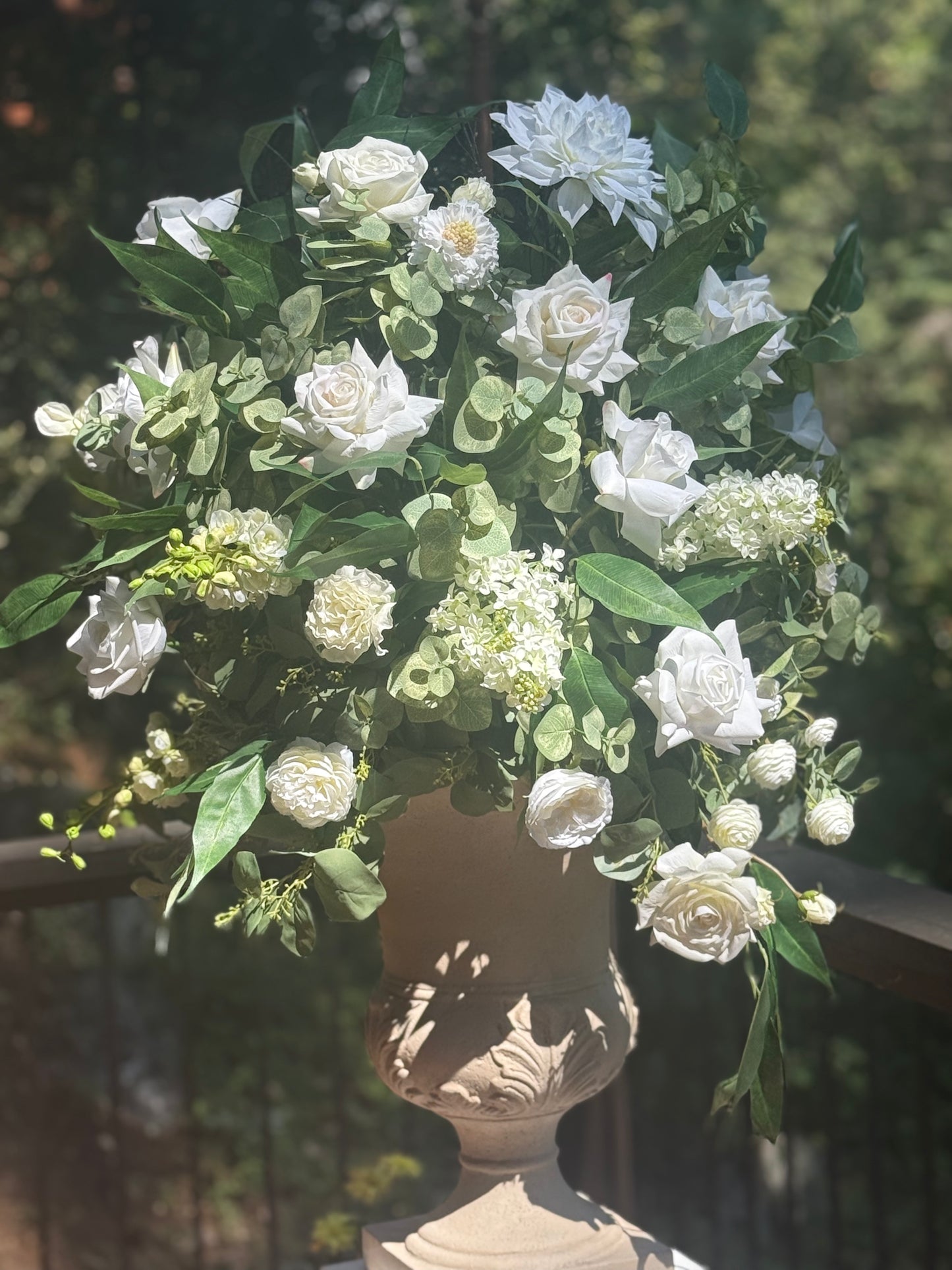 Enchanted floral stone urn rental on wood deck railing with white roses, white dahlias, white hydrangea, white ranunculus, and cascading eucalyptus in dappled sunlight at outdoor pine forest wedding venue | Reno, Lake Tahoe & Sacramento