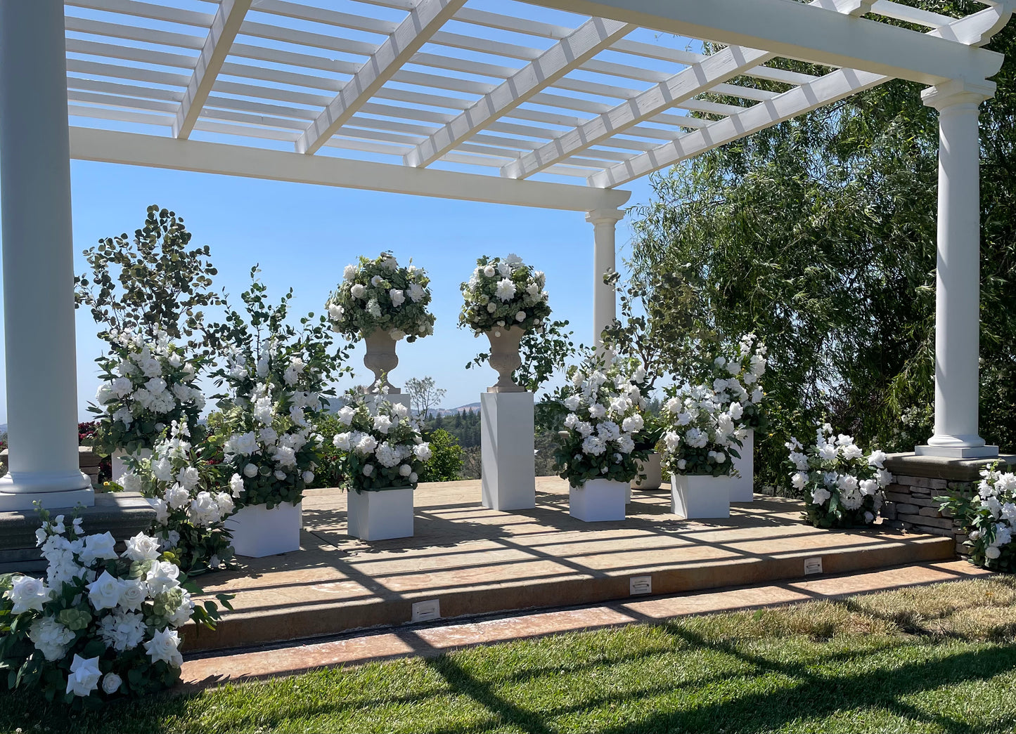 Outdoor wedding ceremony altar under white pergola with Enchanted aisle flower rentals and stone urn rentals on white square pedestals with white roses, white gardenias, and lush eucalyptus along with eucalyptus trees with blue sky and mountain view | Reno, Lake Tahoe & Sacramento