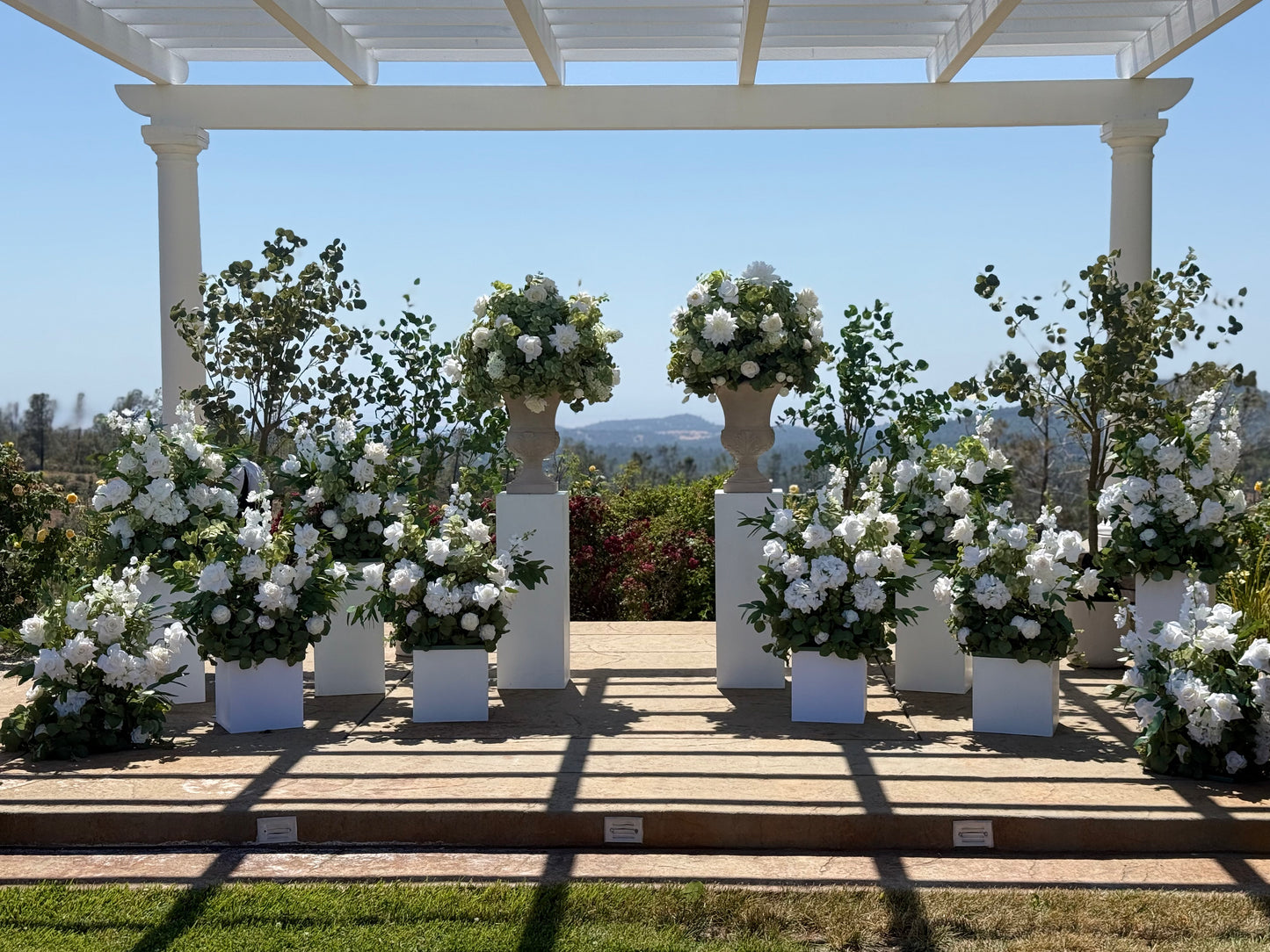 Grand outdoor wedding ceremony setup with two Enchanted floral stone urn rentals on tall white pedestals, Enchanted aisle flowers in white square boxes, and faux eucalyptus trees under white pergola with mountain and blue sky backdrop | Reno, Lake Tahoe & Sacramento