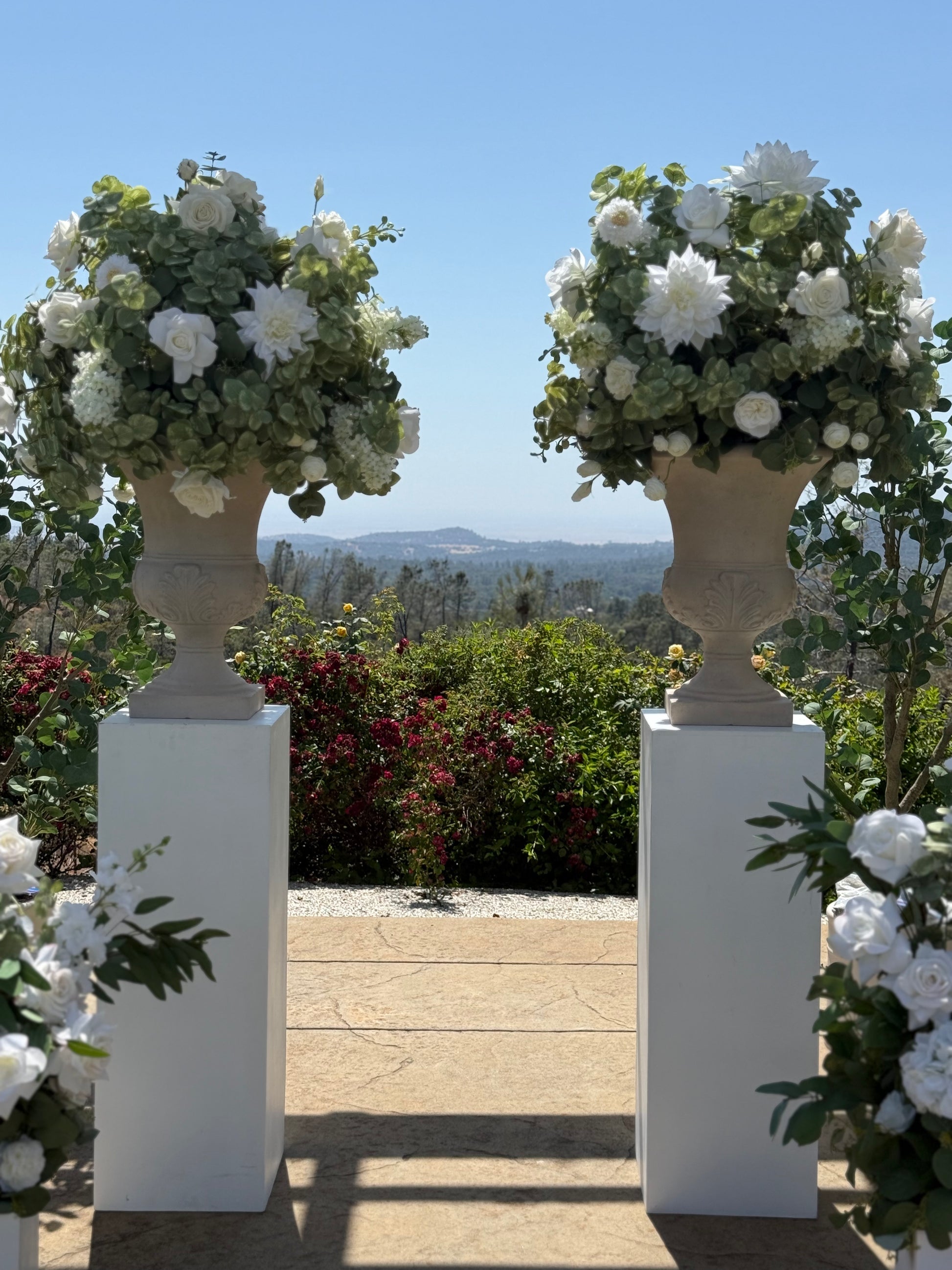 Two Enchanted floral stone urn rentals on white square pedestals framing outdoor stone patio entrance with white roses, white dahlias, and lush greenery with mountain and blue sky backdrop | Reno, Lake Tahoe & Sacramento