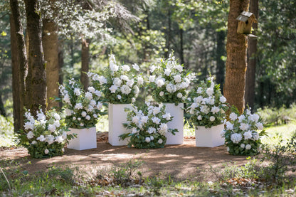 Seven Enchanted aisle flower rentals on white square pedestals with white roses, white gardenias, white delphinium, and lush greenery in dappled sunlight at outdoor pine forest wedding venue | Reno, Lake Tahoe & Sacramento
