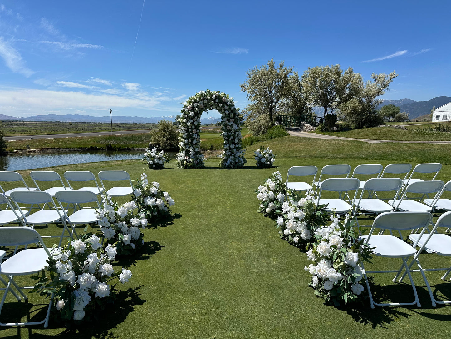 Wide view of Enchanted circular arch rental at outdoor Reno Nevada wedding ceremony with white roses and greenery, Enchanted aisle flowers, mountain and river backdrop, and white folding chairs | Reno, Lake Tahoe & Sacramento