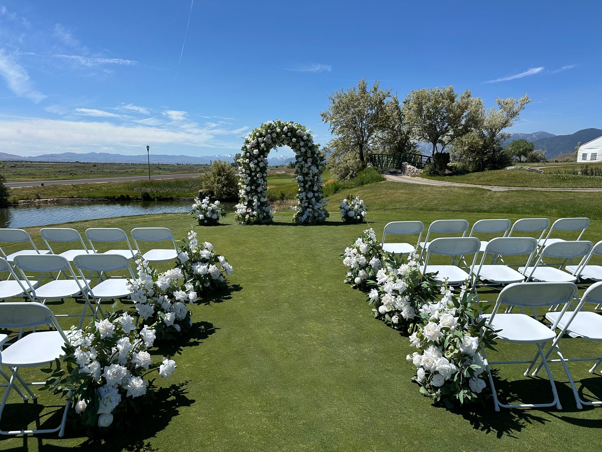 Wide view of Enchanted circular arch rental at outdoor Reno Nevada wedding ceremony with white roses and greenery, Enchanted aisle flowers, mountain and river backdrop, and white folding chairs | Reno, Lake Tahoe & Sacramento