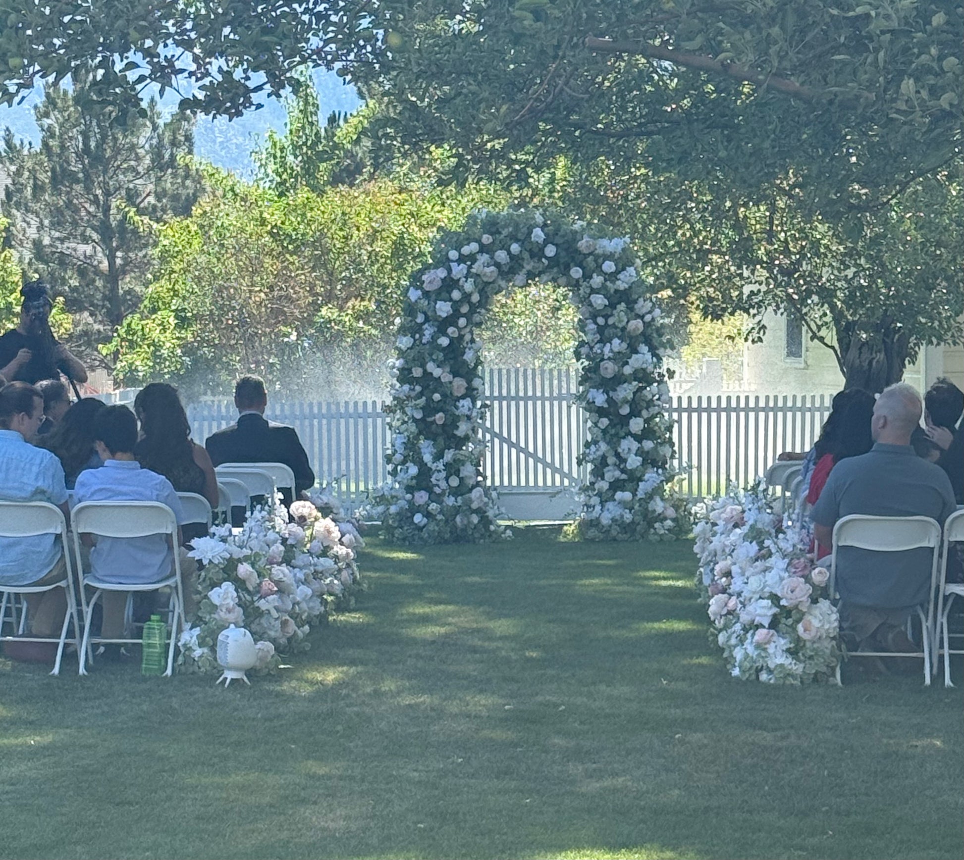 Live wedding ceremony with guests seated as Enchanted circular arch rental with white and peach roses and greenery stands at altar with Love Story aisle flowers lining outdoor garden venue | Reno, Lake Tahoe & Sacramento