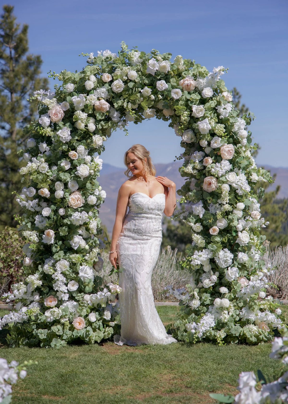 Bride standing under Enchanted circular arch rental with white roses, blush peonies, and greenery at outdoor Reno Nevada wedding venue and mountain backdrop | Reno, Lake Tahoe & Sacramento