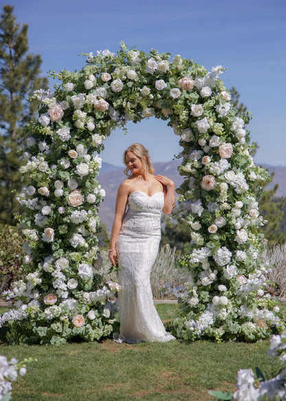 Bride standing under Enchanted circular arch rental with white roses, blush peonies, and greenery at outdoor Reno Nevada wedding venue and mountain backdrop | Reno, Lake Tahoe & Sacramento