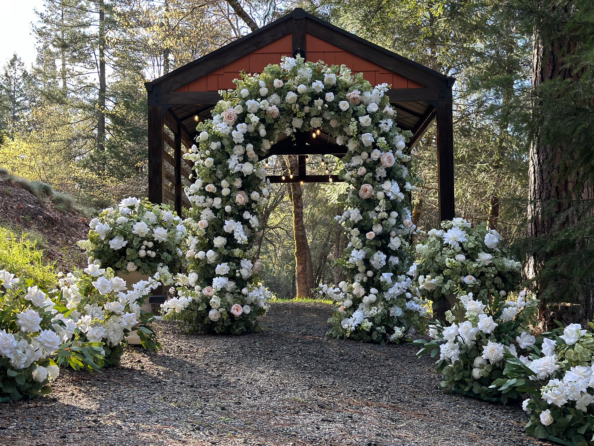 Enchanted circular arch rental with white and blush roses and greenery in front of wood pavilion at outdoor pine forest wedding venue with Enchanted aisle flowers and urns | Reno, Lake Tahoe & Sacramento