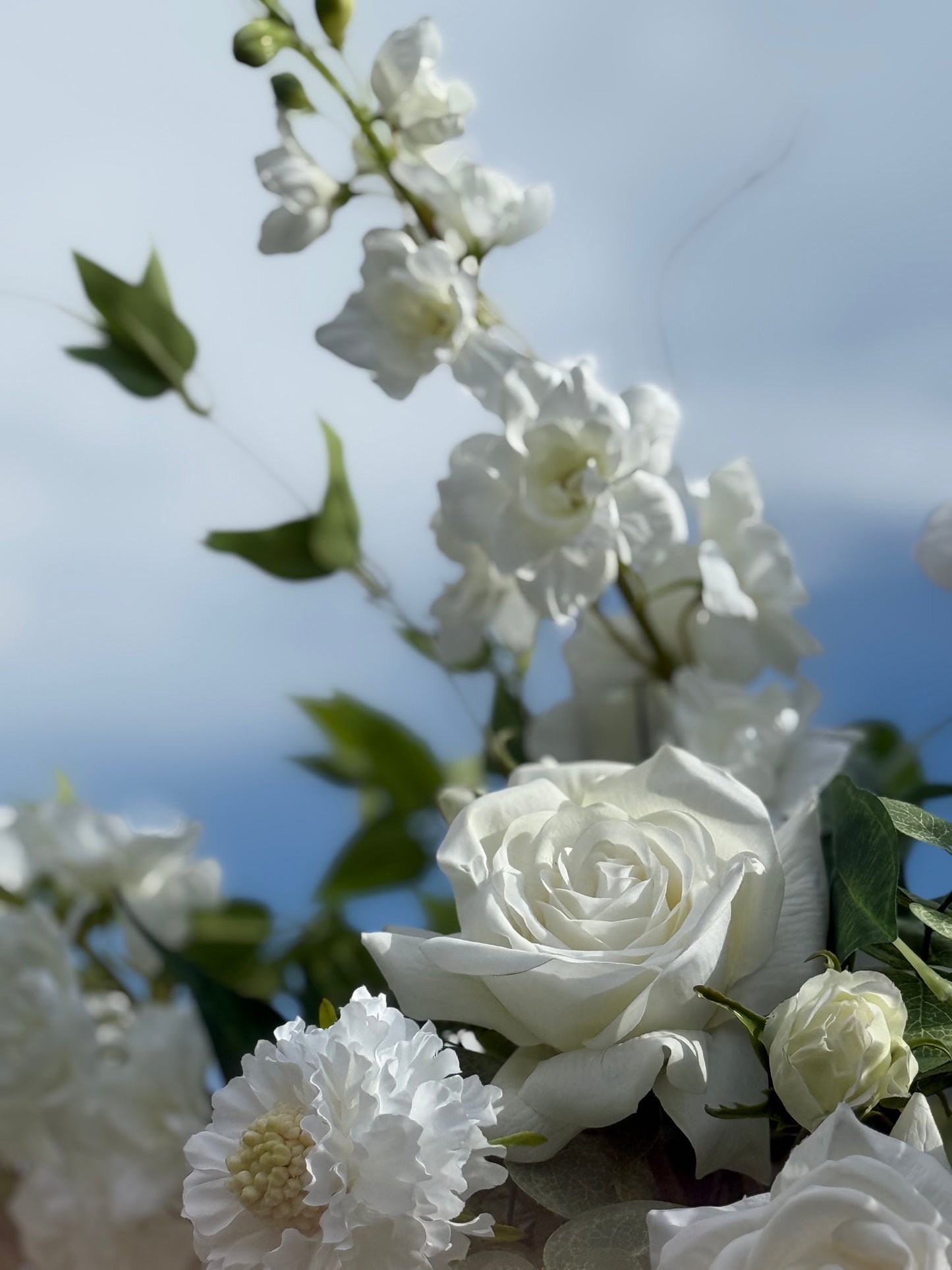 Close-up of Enchanted floral urn rental blooms with white rose, white delphinium, white dahlias, and white ranunculus buds against bright blue sky at outdoor wedding venue | Reno, Lake Tahoe & Sacramento