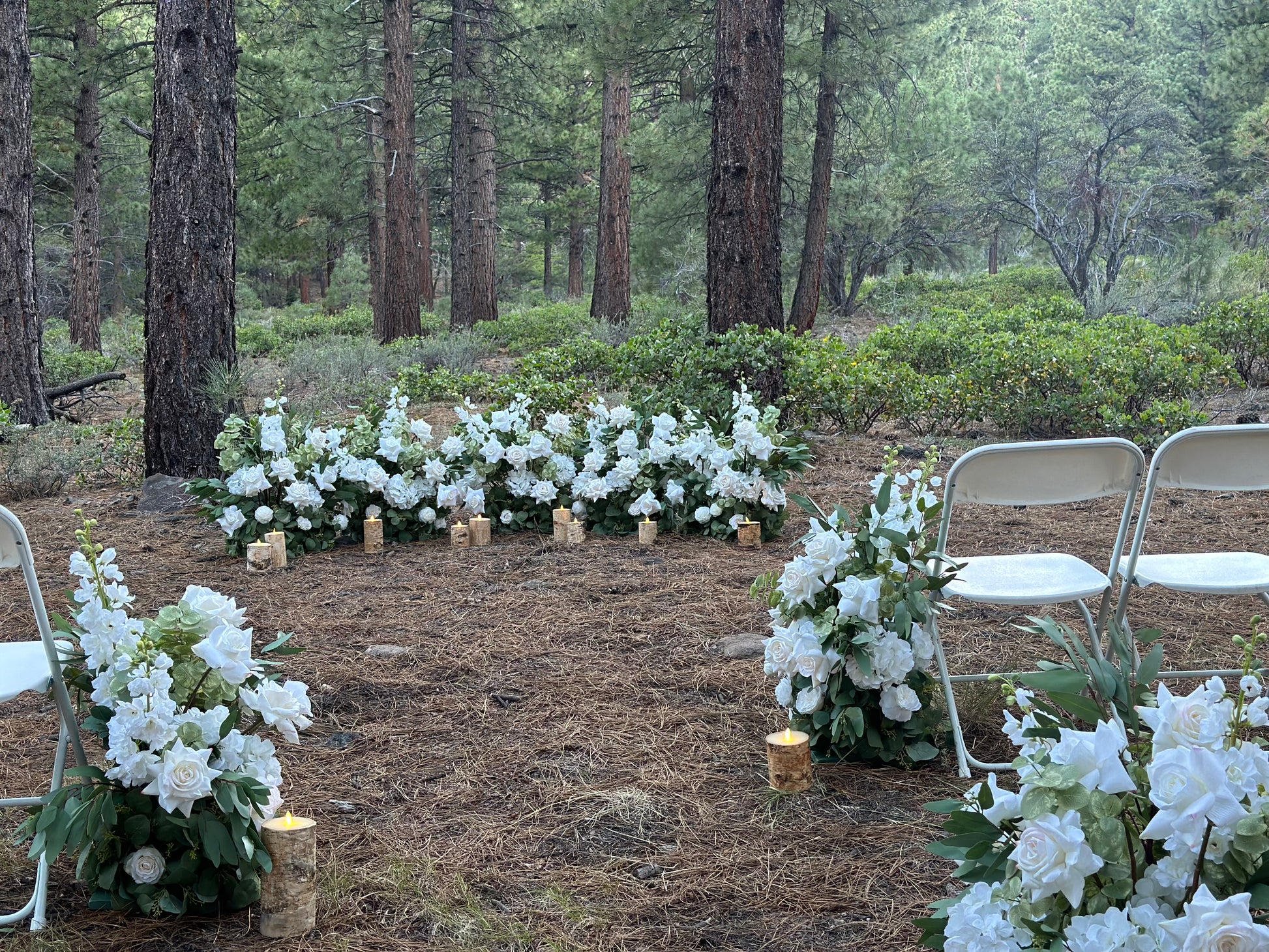 Enchanted aisle flowers designed in meadow hedge shape lining outdoor pine forest wedding ceremony aisle with white roses, white hydrangea, white gardenias, lush greenery, and birch wood candles beside white folding chairs | Reno, Lake Tahoe & Sacramento