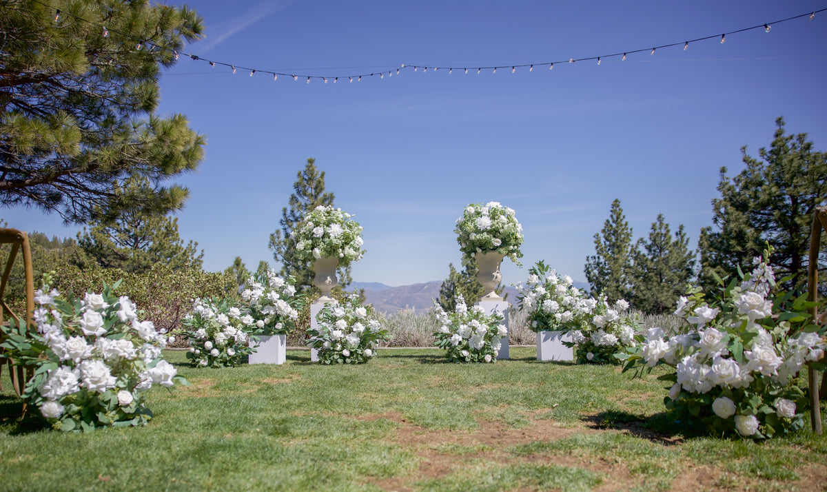 Wide view of Enchanted aisle flower rentals and urn rentals on white pedestals lining outdoor mountain wedding ceremony aisle with pine trees, blue sky, and string lights | Reno, Lake Tahoe & Sacramento