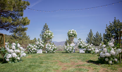 Wide view of Enchanted aisle flower rentals and urn rentals on white pedestals lining outdoor mountain wedding ceremony aisle with pine trees, blue sky, and string lights | Reno, Lake Tahoe & Sacramento