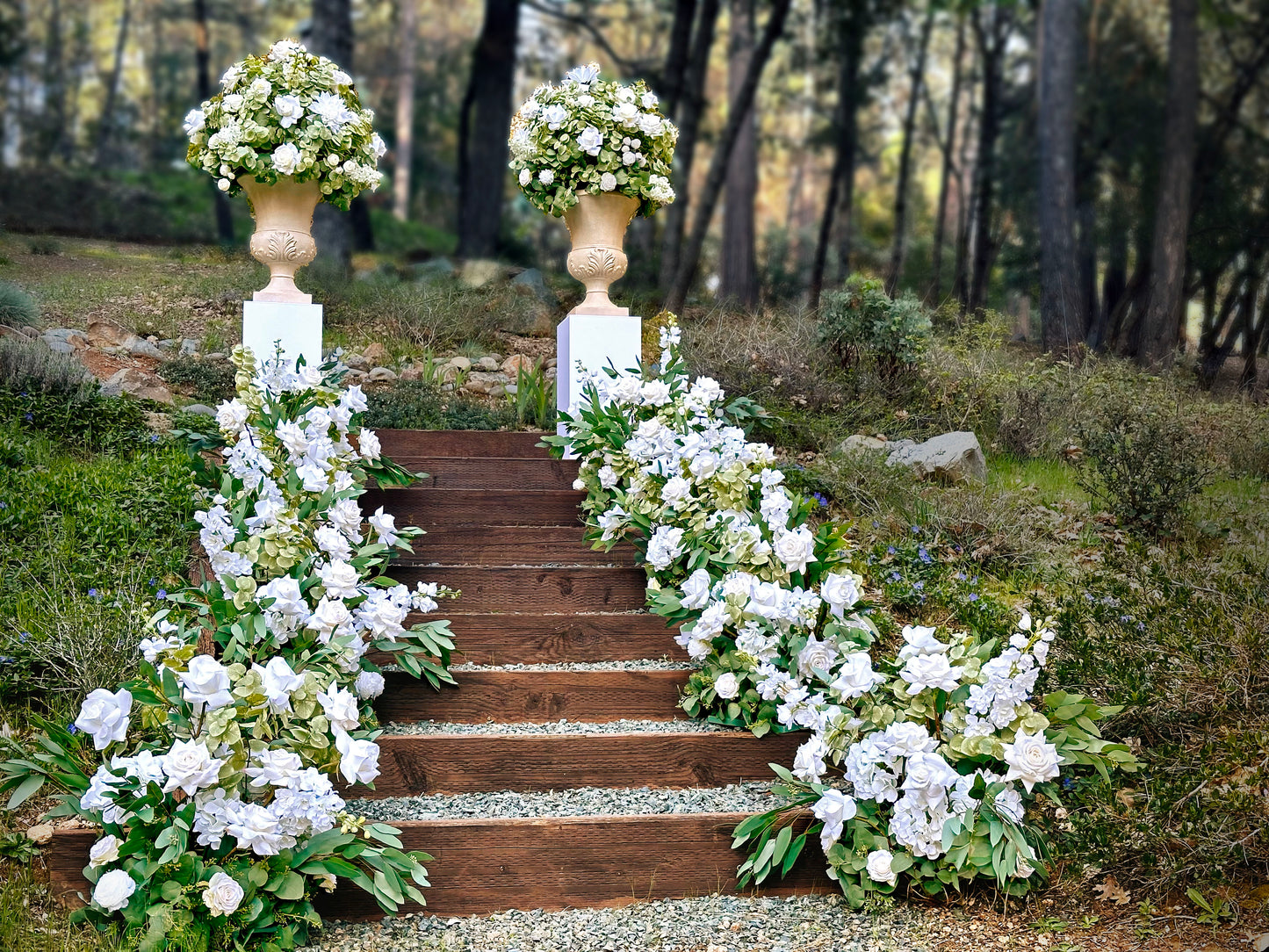 Two stone urn rentals on white pedestals flanking wood staircase lined with Enchanted aisle flower rentals cascading with white roses, white hydrangea, white gardenias, and lush greenery at outdoor pine forest wedding venue | Reno, Lake Tahoe & Sacramento