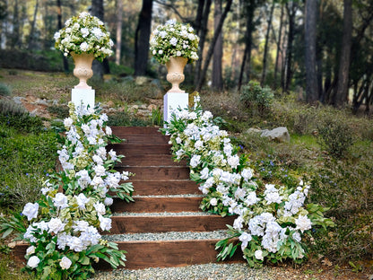 Two stone urn rentals on white pedestals flanking wood staircase lined with Enchanted aisle flower rentals cascading with white roses, white hydrangea, white gardenias, and lush greenery at outdoor pine forest wedding venue | Reno, Lake Tahoe & Sacramento