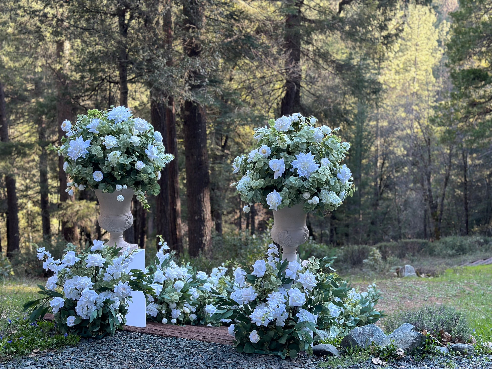Two Enchanted floral stone urn rentals on white pedestals with Enchanted aisle flower rentals cascading at base with white roses, white dahlias, white delphinium, and lush greenery at outdoor pine forest wedding venue | Reno, Lake Tahoe & Sacramento
