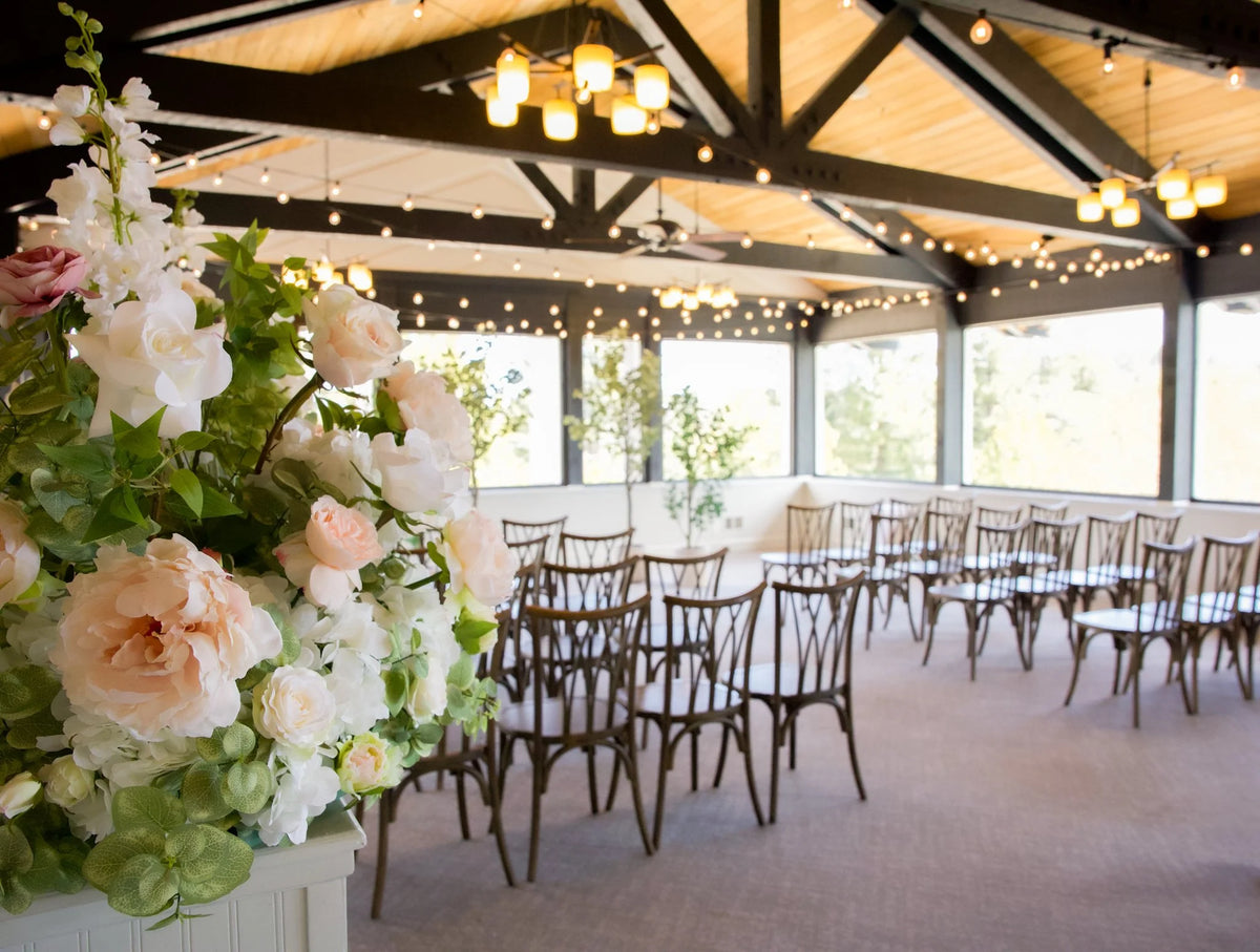 Love Story aisle flower rental with blush roses and white peonies on white counter in foreground with faux eucalyptus tree rentals, cross-back chairs, and string lights at indoor wedding venue | Reno, Lake Tahoe & Sacramento