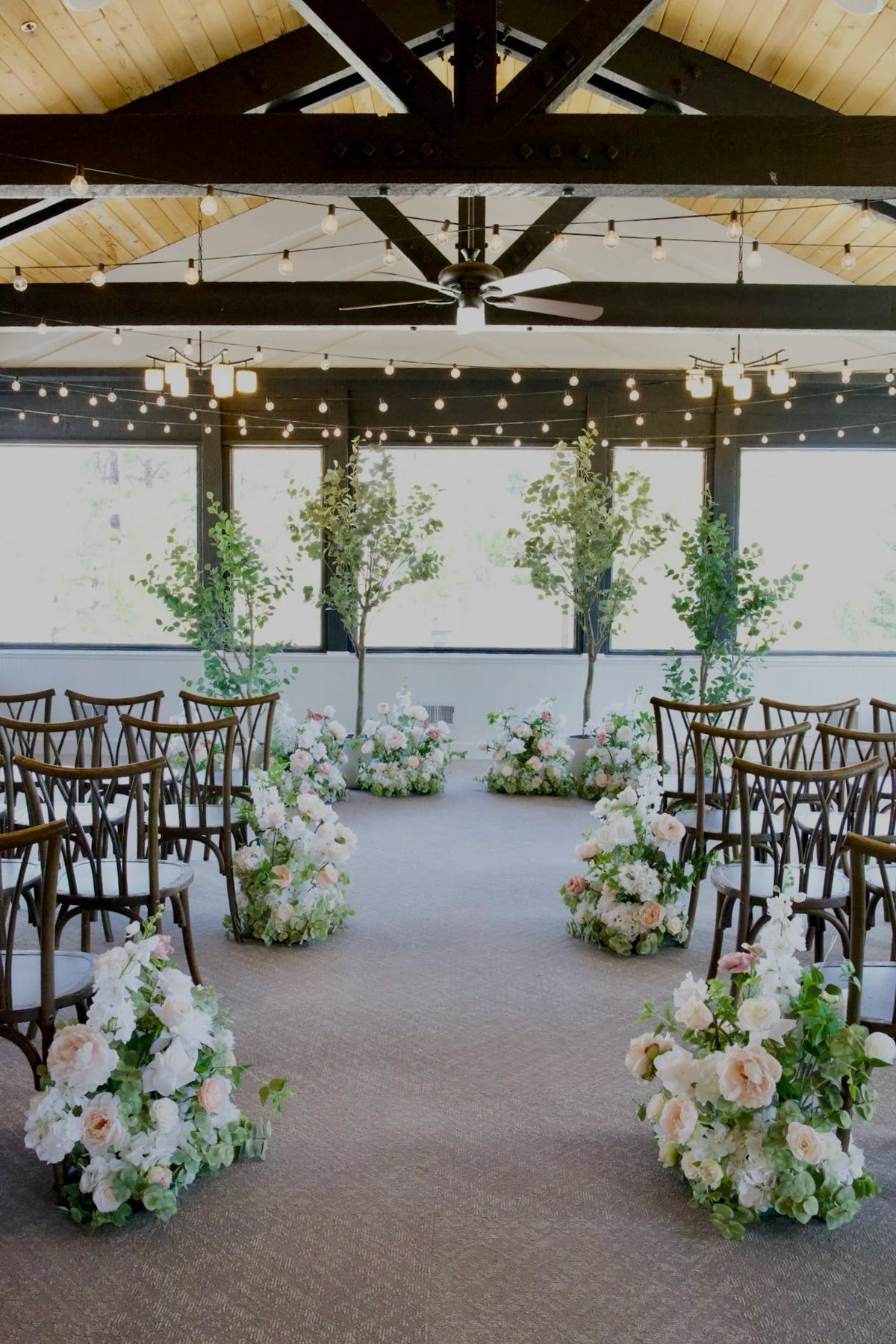 Four faux eucalyptus tree rentals in white cylinder pots with Love Story aisle flowers in blush and white, cross-back chairs, and string lights at Lake Tahoe venue | Reno, Lake Tahoe & Sacramento