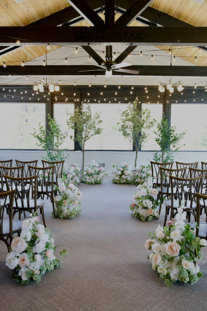 Four faux eucalyptus tree rentals in white cylinder pots with Love Story aisle flowers in blush and white, cross-back chairs, and string lights at Lake Tahoe venue | Reno, Lake Tahoe & Sacramento