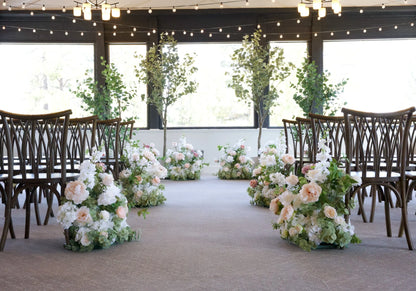 Four faux eucalyptus tree rentals in white cylinder pots flanking indoor Love Story aisle flowers with blush and white ground florals, cross-back chairs, and string lights at Lake Tahoe venue | Reno, Lake Tahoe & Sacramento