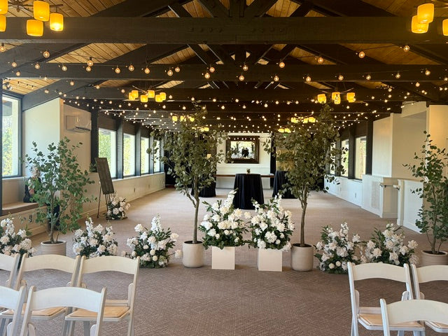 Full room view of faux eucalyptus tree rentals and Enchanted ground florals styled as a room divider separating wedding ceremony seating from cocktail area, white folding chairs, and string lights at indoor venue | Reno, Lake Tahoe & Sacramento
