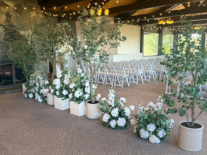 Back view of faux eucalyptus tree rentals and Enchanted aisle flower rentals in white cylinder and square pots lining indoor wedding ceremony aisle with stone fireplace, white folding chairs, and string lights used as room divider | Reno, Lake Tahoe & Sacramento