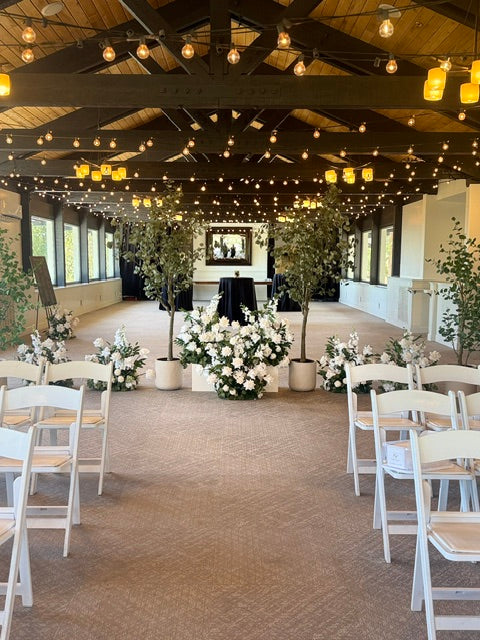 Wide aisle view of faux eucalyptus tree rentals and Enchanted ground florals used as a room divider between wedding ceremony and cocktail area, white folding chairs, and string lights at indoor venue | Reno, Lake Tahoe & Sacramento