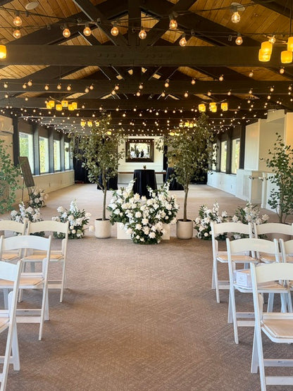 Wide aisle view of faux eucalyptus tree rentals and Enchanted ground florals used as a room divider between wedding ceremony and cocktail area, white folding chairs, and string lights at indoor venue | Reno, Lake Tahoe & Sacramento