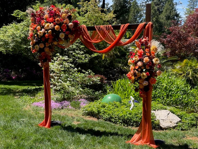Full Evermore Fall arch adornment rental with coral roses, orange ranunculus, burgundy dahlias, and rust chiffon drapery at outdoor garden wedding venue with lush greenery backdrop | Reno, Lake Tahoe & Sacramento