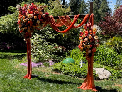 Full Evermore Fall arch adornment rental with coral roses, orange ranunculus, burgundy dahlias, and rust chiffon drapery at outdoor garden wedding venue with lush greenery backdrop | Reno, Lake Tahoe & Sacramento